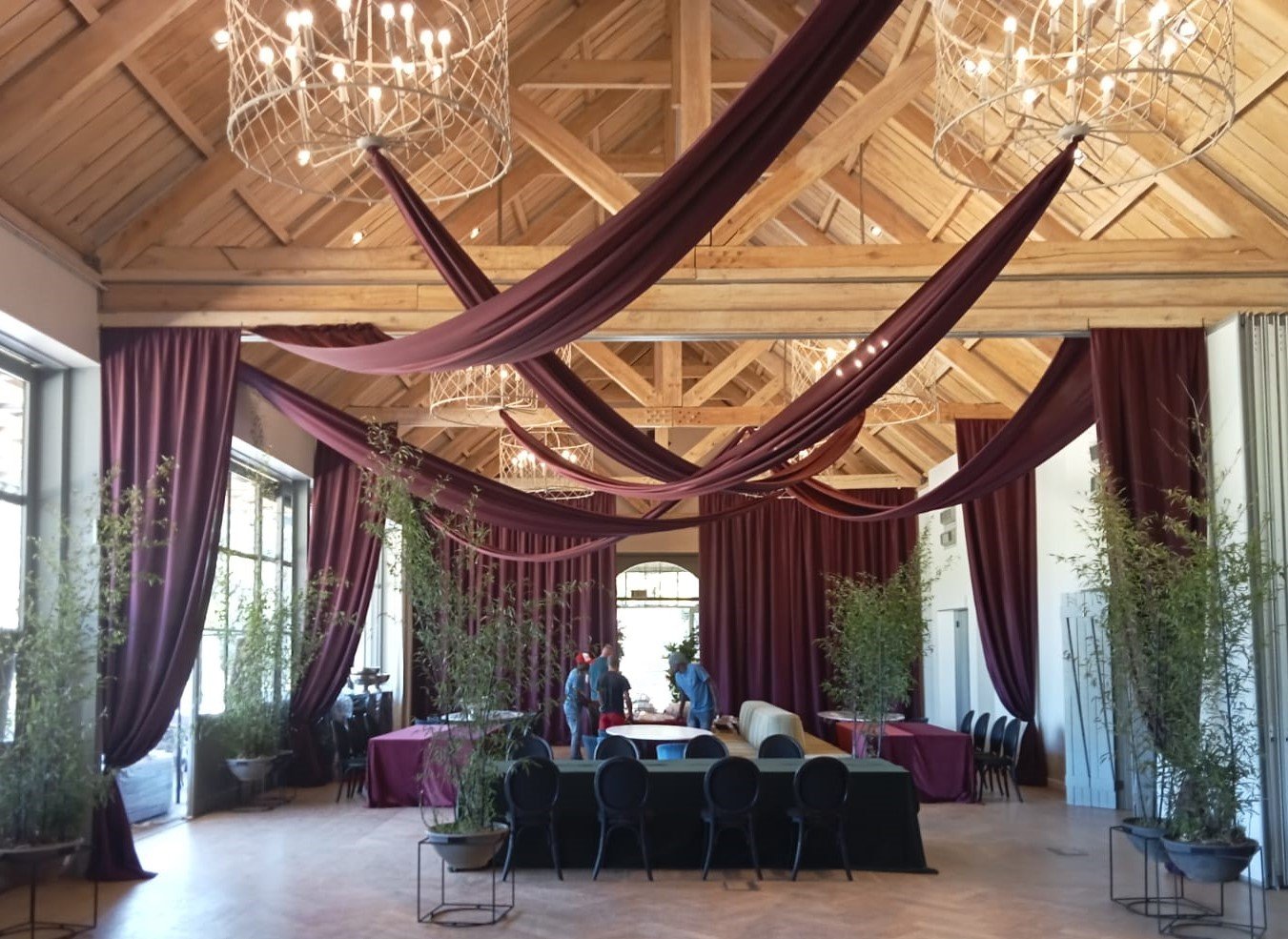 Burgundy random looping ceiling swags with a series of side-facing tieback draping swags, complemented by chandeliers and greenery, for an elegant indoor event by The Events Draping Co.