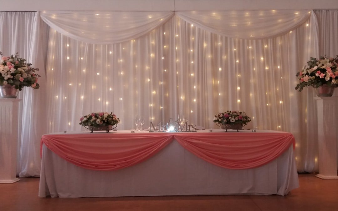 Bridal backdrop at Joostenberg Wine Estate, Stellenbosch, featuring white fabric draping with swags and fairy lights, and peach fabric swags across the bridal table by The Events Draping Co.