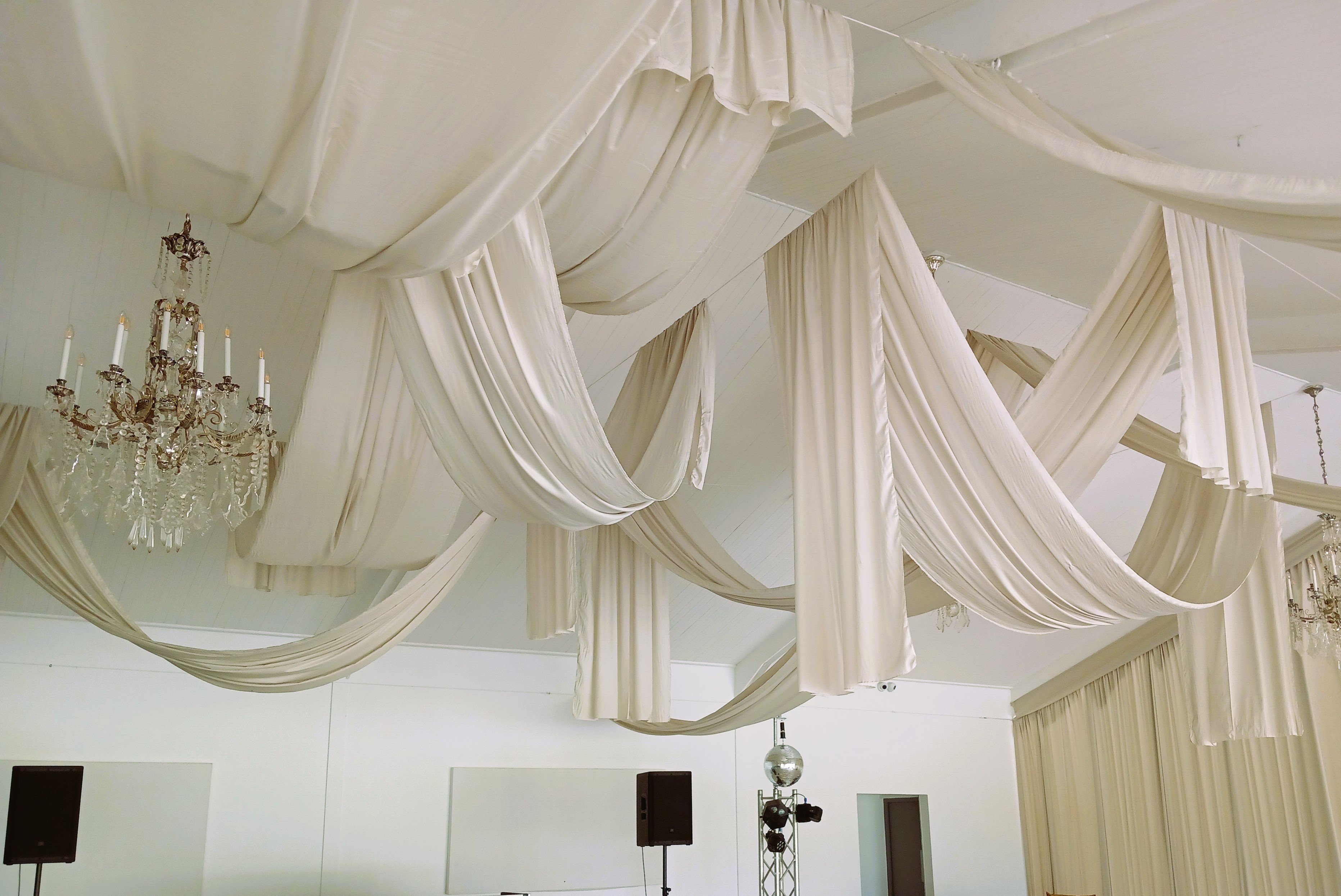 Random ceiling swags with drops in cream fabric, complemented by chandeliers, creating an elegant and sophisticated look for an indoor event by The Events Draping Co.