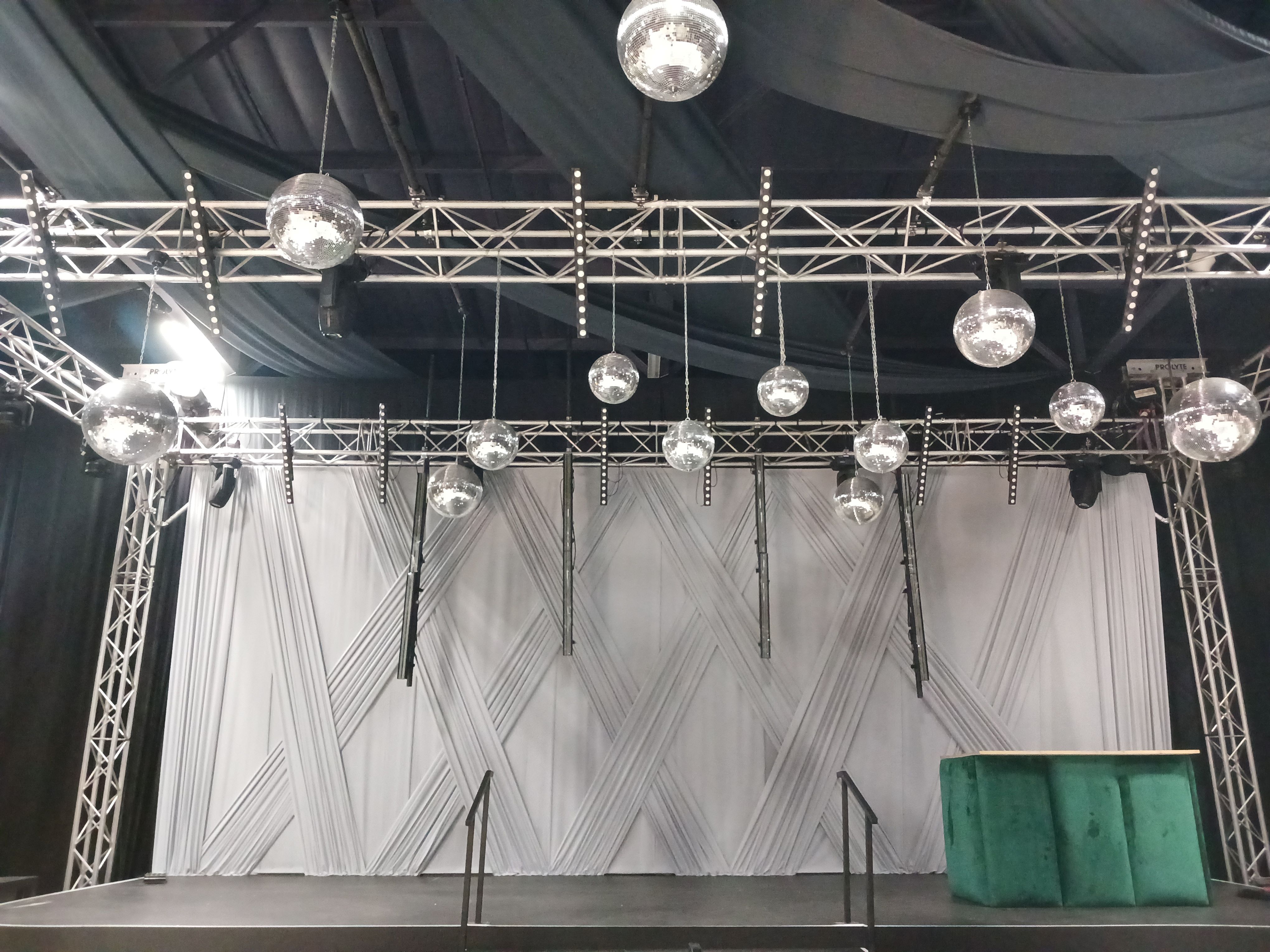 Large criss-cross stage backdrop, 8 meters high by 14 meters wide, in light grey fabric, featuring hanging disco balls for an event setup by The Events Draping Co