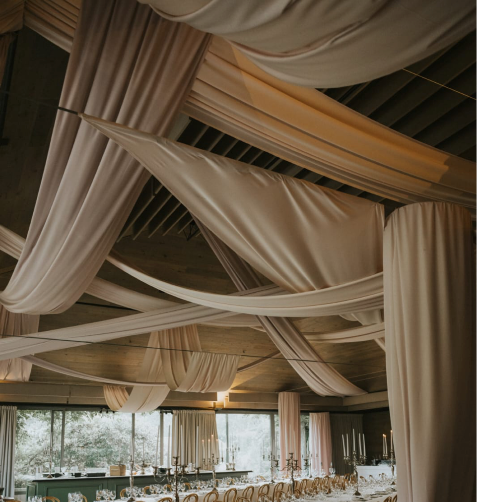 Elegant indoor event setup with random ceiling swags in beige fabric, creating a sophisticated atmosphere, complemented by candlelit dining tables, by The Events Draping Co.
